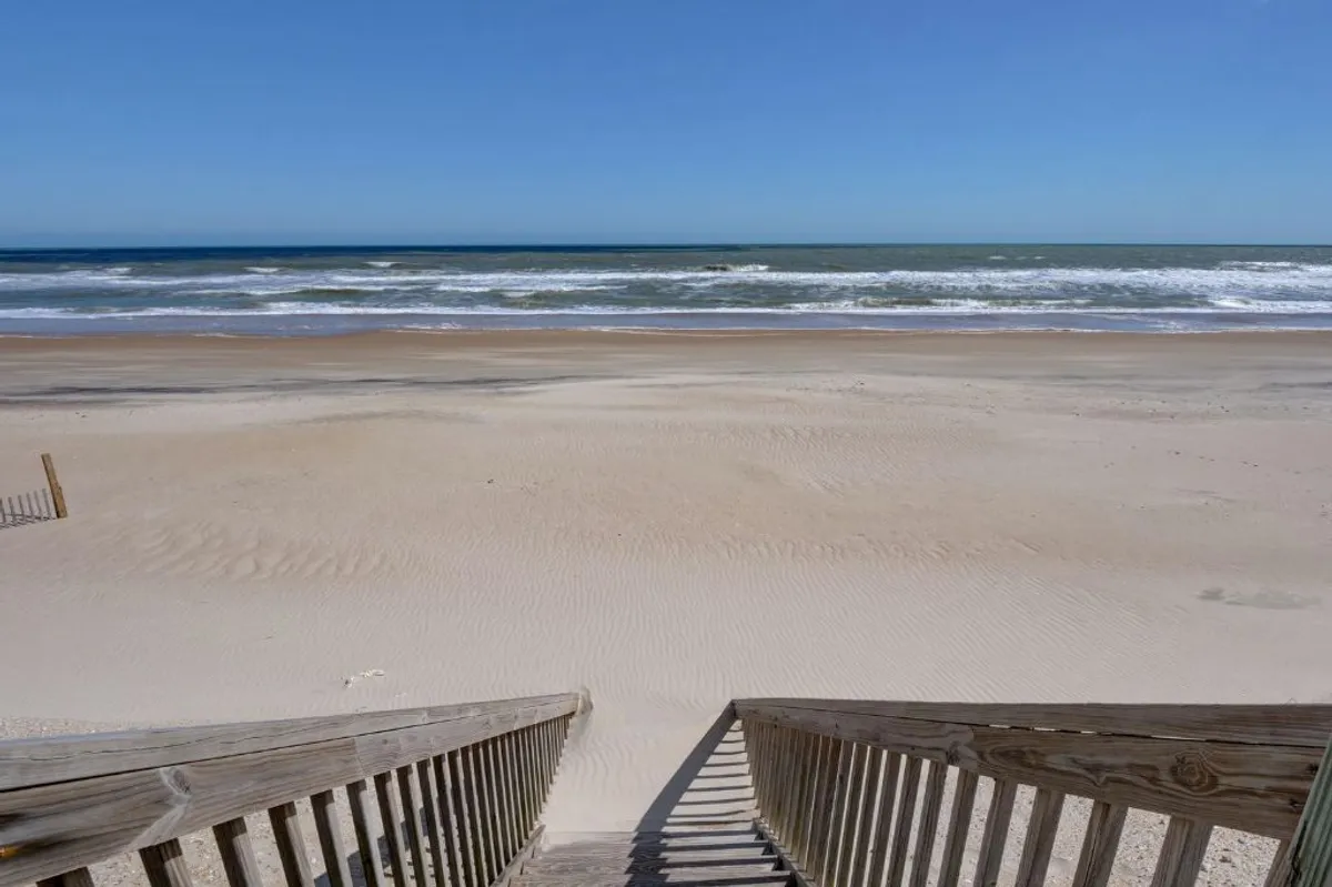 A wide view of the vast sandy beach bordered by gentle ocean waves, with wooden stairs providing private, direct access from the property. The clear blue sky suggests a perfect day for family activities by the sea.