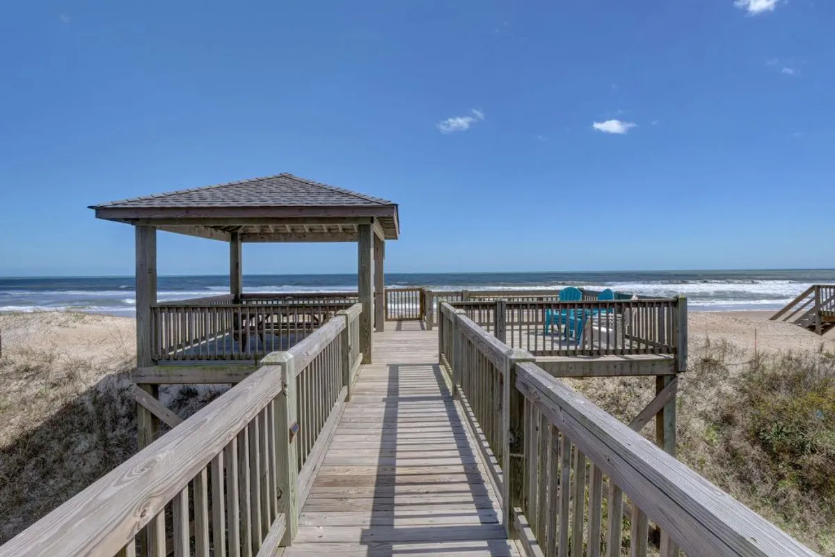 An inviting exterior scene showcasing a wooden boardwalk and a gazebo overlooking the ocean, with lounge chairs and an open view of the beach. The area invites gatherings, relaxation, and socializing as the sun rises or sets over the water.