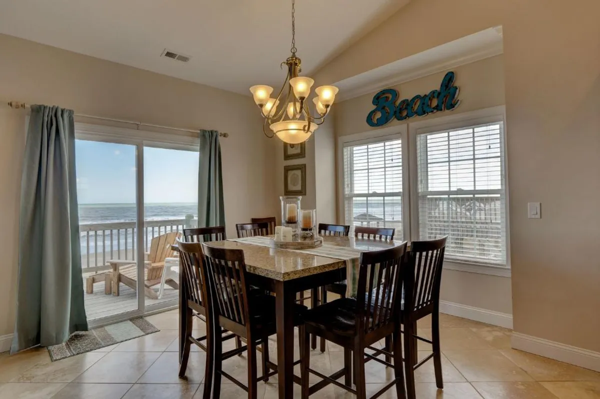 A cheerful interior dining space features a large table set beside windows offering a clear beach view. The atmosphere is bright and welcoming, ideal for group meals, celebrations, and reconnecting over local seafood feasts.