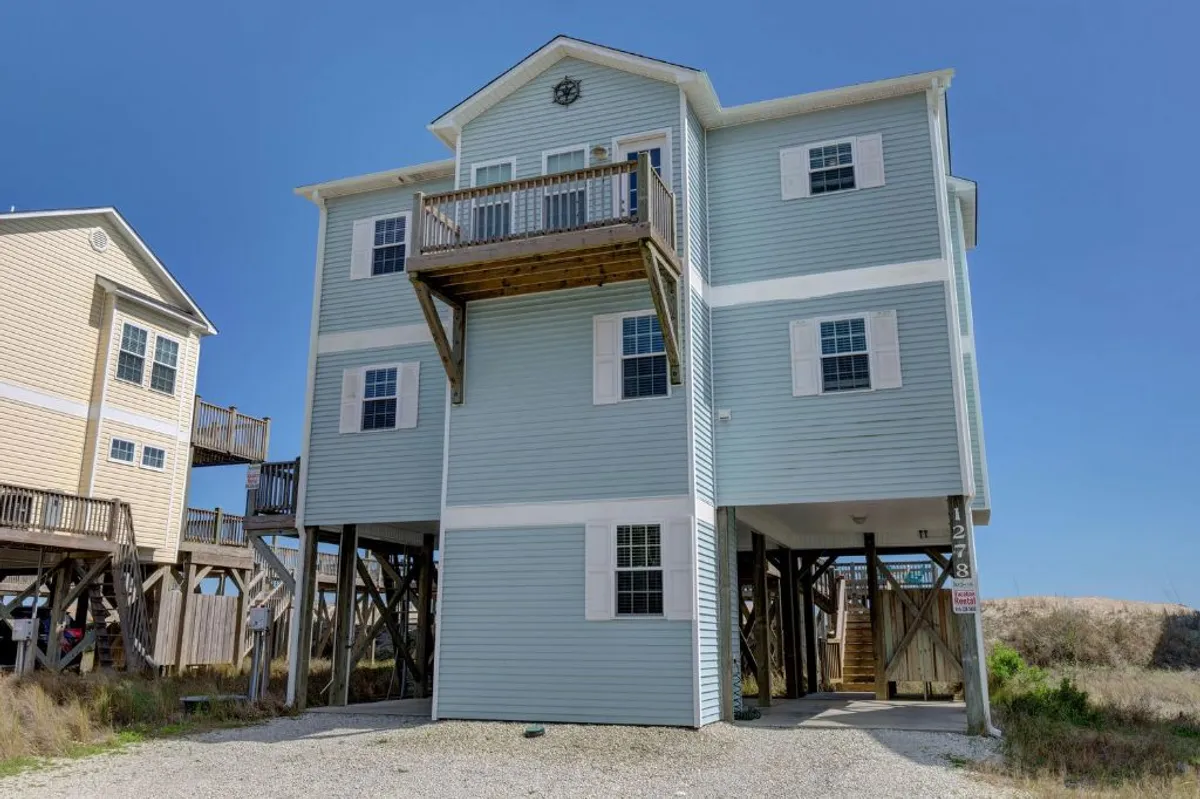 The image captures the exterior of the home, showcasing its elevated deck, light blue siding, and stilted foundation. Ample space is visible for both covered and open parking, making it clear that arrivals and departures are easy for guests with vehicles. The deck also hints at comfortable outdoor living and convenient access to the home.