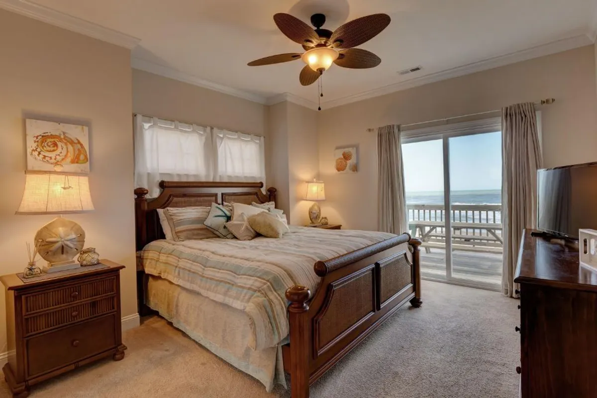 A tranquil bedroom featuring a bed positioned beside a sliding glass door that opens directly to an outdoor deck. Nightstands and a ceiling fan add comfort, while the proximity to the outdoors highlights the seamless transition between private space and the beachside environment.