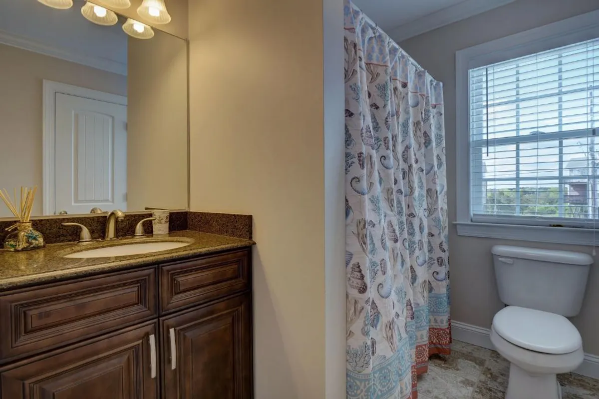 A calming bathroom featuring a granite countertop, decorative shower curtain, and a well-lit vanity mirror. The atmosphere is serene and inviting, ideal for relaxing and refreshing after a day at the beach.