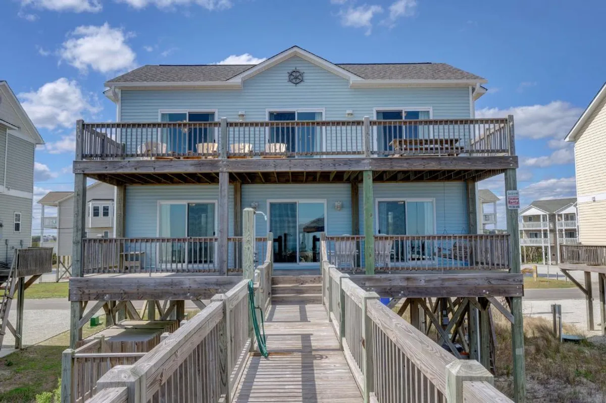 The image showcases the home's exterior, highlighting a spacious two-story wooden deck adorned with comfortable outdoor furniture and charming nautical décor. Large glass doors invite the outdoors in, while the deck offers panoramic sunset views over the North Topsail Beach shoreline. The mood feels casual and inviting, perfect for family gatherings or relaxed evenings overlooking the ocean.