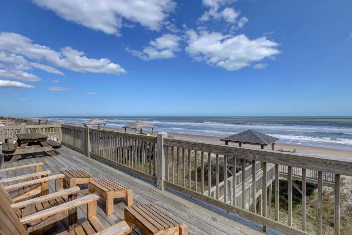 This image features a spacious wooden deck overlooking the ocean under a cloudy sky. The deck is furnished for relaxation and gatherings, with ample space for seating, and offers unobstructed views of the water. The tranquil atmosphere and open-air setting capture the essence of oceanfront outdoor living.