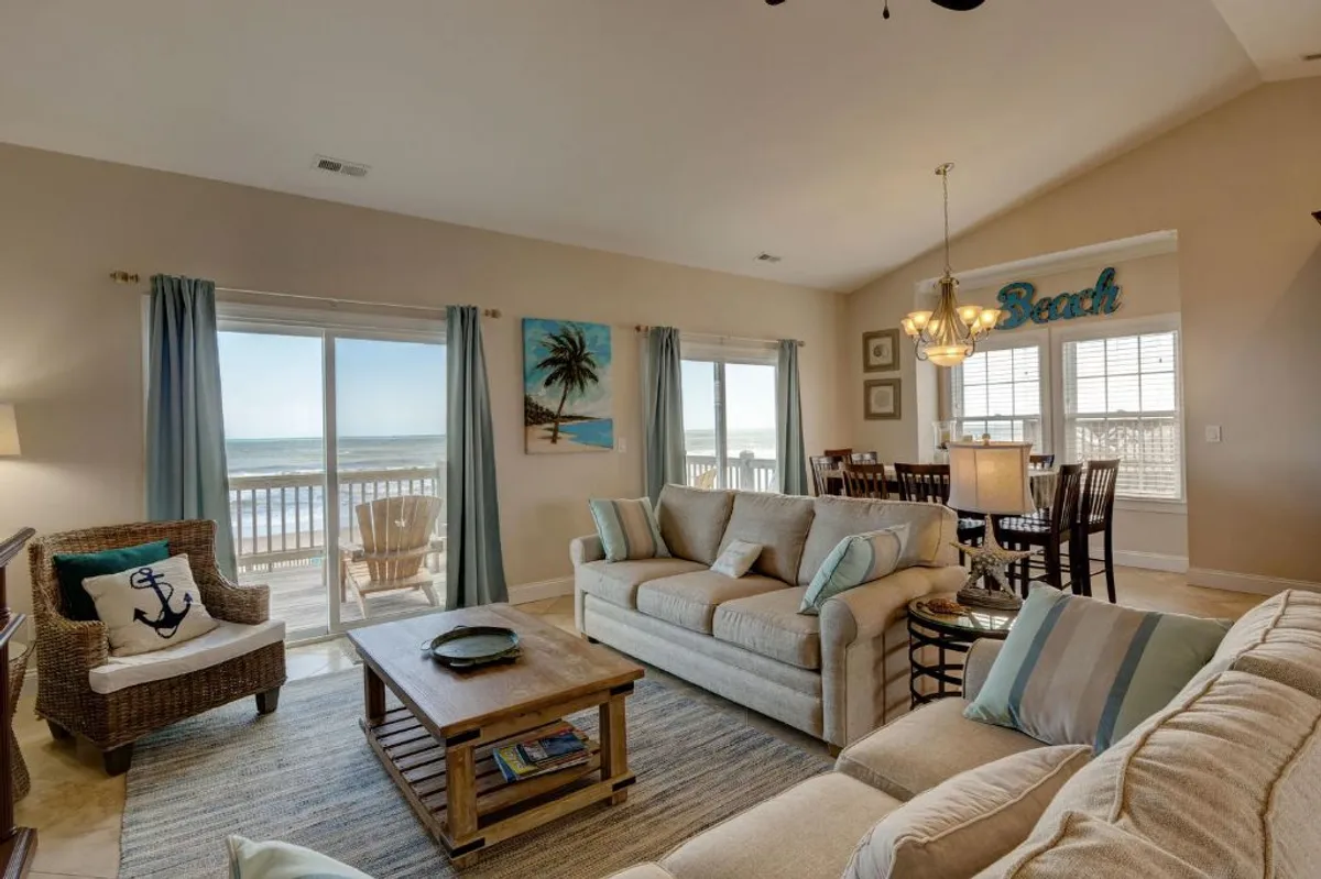This image showcases a bright, open-plan living and dining area with large windows framing stunning ocean views. The space features a comfortable sofa and a dining area adjacent to the windows, with beach-themed artwork lending a breezy, relaxed vibe.