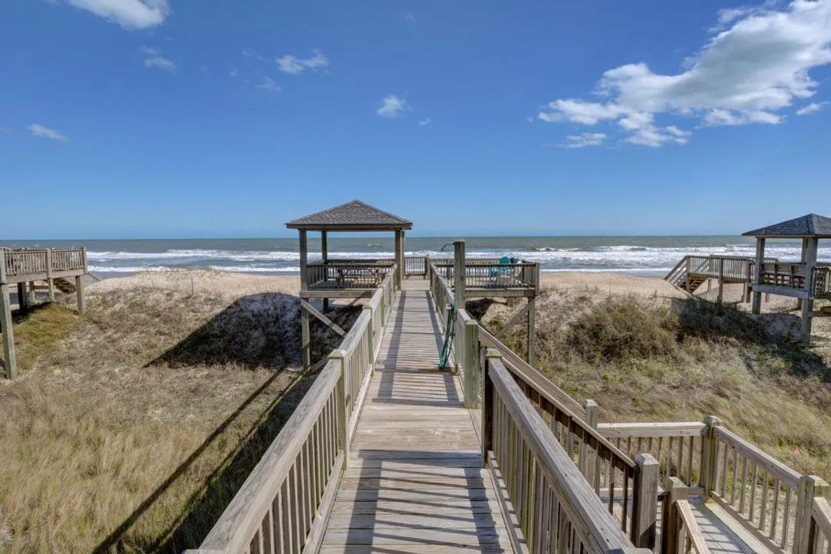 This exterior image features a wooden boardwalk leading through dune grass to a sandy beach, with gentle ocean waves in the background. Beach huts dot the shoreline, inviting relaxation and group activities by the coast.