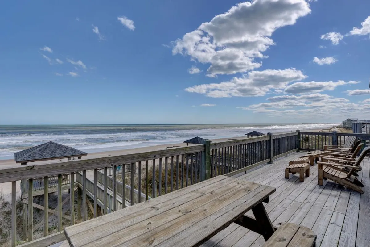 A wooden deck with scenic ocean views, complemented by beachside structures and a peaceful, cloudy sky. The deck is an inviting space for activities like sunbathing or enjoying morning coffee while taking in the ocean breeze. The beach is visible just steps away, emphasizing easy access.