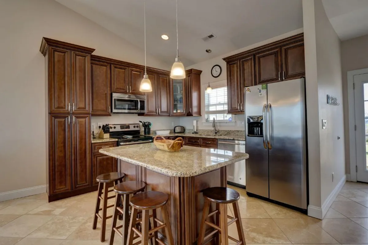 A modern kitchen with granite countertops, wood cabinetry, stainless steel appliances, and island seating is prominent in this image. The space feels warm and functional, perfect for guests seeking convenience during their stay. The kitchen's appeal is enhanced by details that imply ease of use and professional upkeep.
