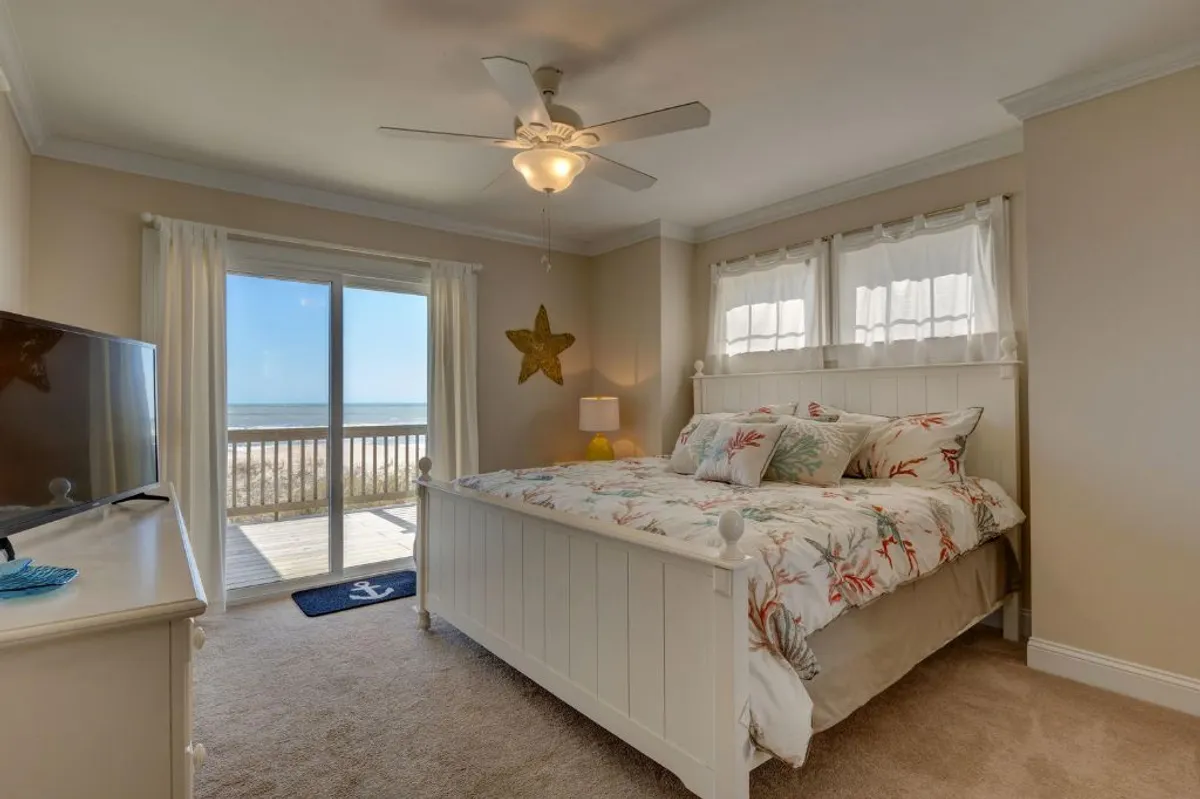 A tranquil bedroom featuring a bed with floral bedding, a large window offering sweeping ocean views, starfish decorations, and a television, all bathed in natural light. The room’s decor is bright and beach-inspired, creating a restful, airy sanctuary.